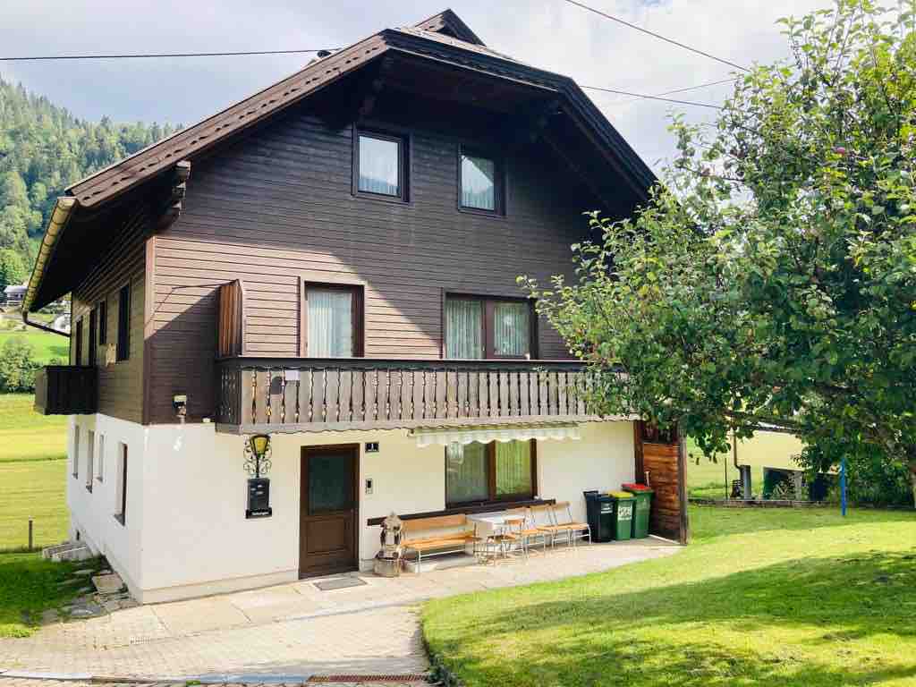 A charming two-story wooden house is set against a backdrop of lush greenery. The exterior features a covered balcony with seating and large windows that allow natural light to enter. The well-maintained garden area includes a path leading to the main entrance.
