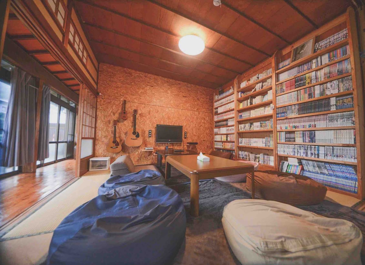 A warm living space features wooden walls and flooring, complemented by bookshelves filled with numerous manga. Comfortable bean bags are arranged around a central low table, with guitars displayed nearby. A television is positioned in front, offering a cozy environment for relaxation.