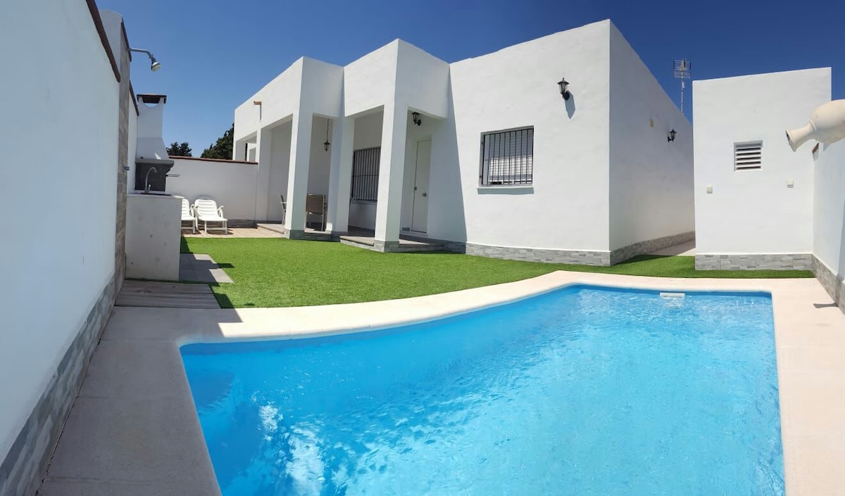 A private area showcases a clear blue swimming pool surrounded by artificial grass. The white-walled structure features multiple entrances and is accented by two lounge chairs. Sunlight streams onto the scene, highlighting the inviting outdoor space.