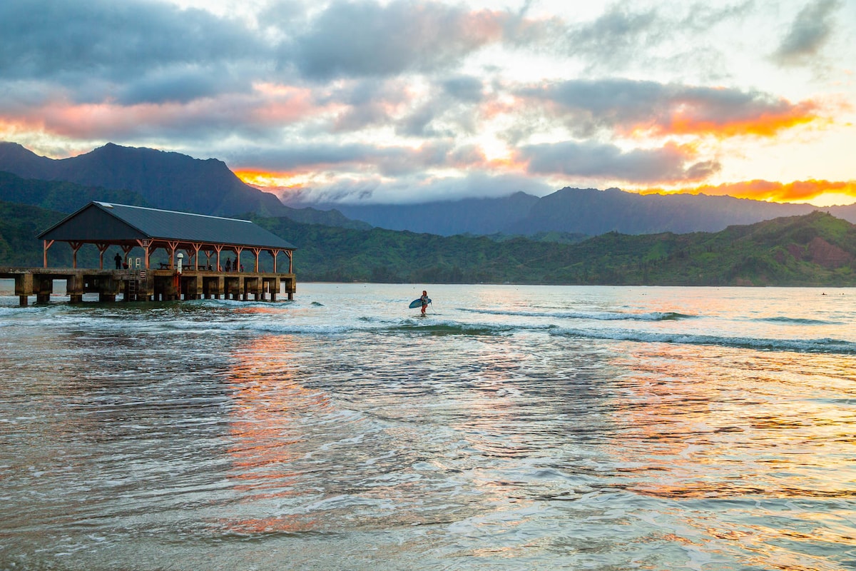 A tranquil scene depicts a lone surfer riding gentle waves near a wooden pier. The backdrop features lush mountains under a twilight sky painted with soft hues of orange and gray. The calm water reflects the serene atmosphere of the Hanalei Bay.