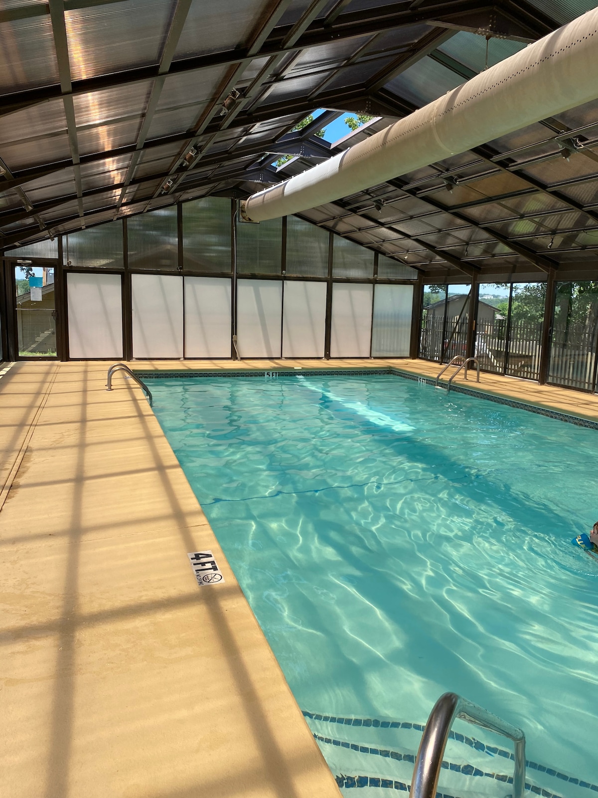 An indoor pool is viewed under a skylight, allowing natural light to illuminate the clear water. The smooth deck surrounds the pool, and the space is enclosed with glass windows, providing a serene atmosphere for relaxation. Visibility of lane markers is present on the pool floor.