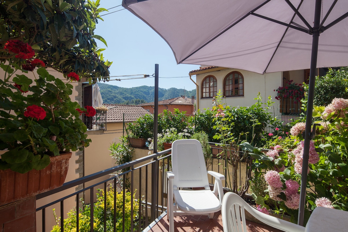 The terrace is adorned with vibrant flowers and greenery. A white chair is positioned under a light purple umbrella, providing shade. Surrounding the space are various planters filled with blooming plants, while distant hills are visible in the background against a clear blue sky.