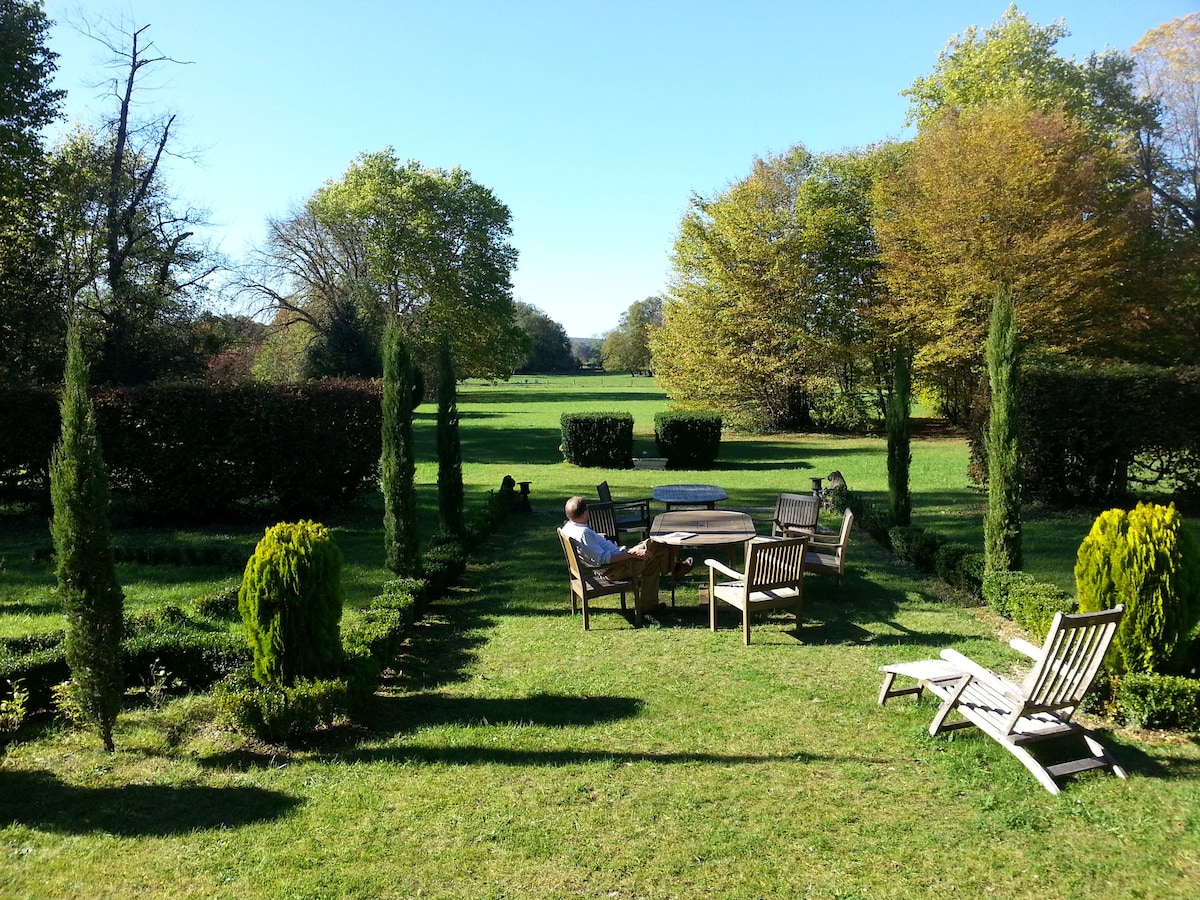 A serene outdoor seating area is featured, surrounded by neatly trimmed hedges and trees, set against a lush green lawn. Wooden chairs are positioned around a circular table, while a pair of loungers can be seen nearby, inviting relaxation in a peaceful landscape.