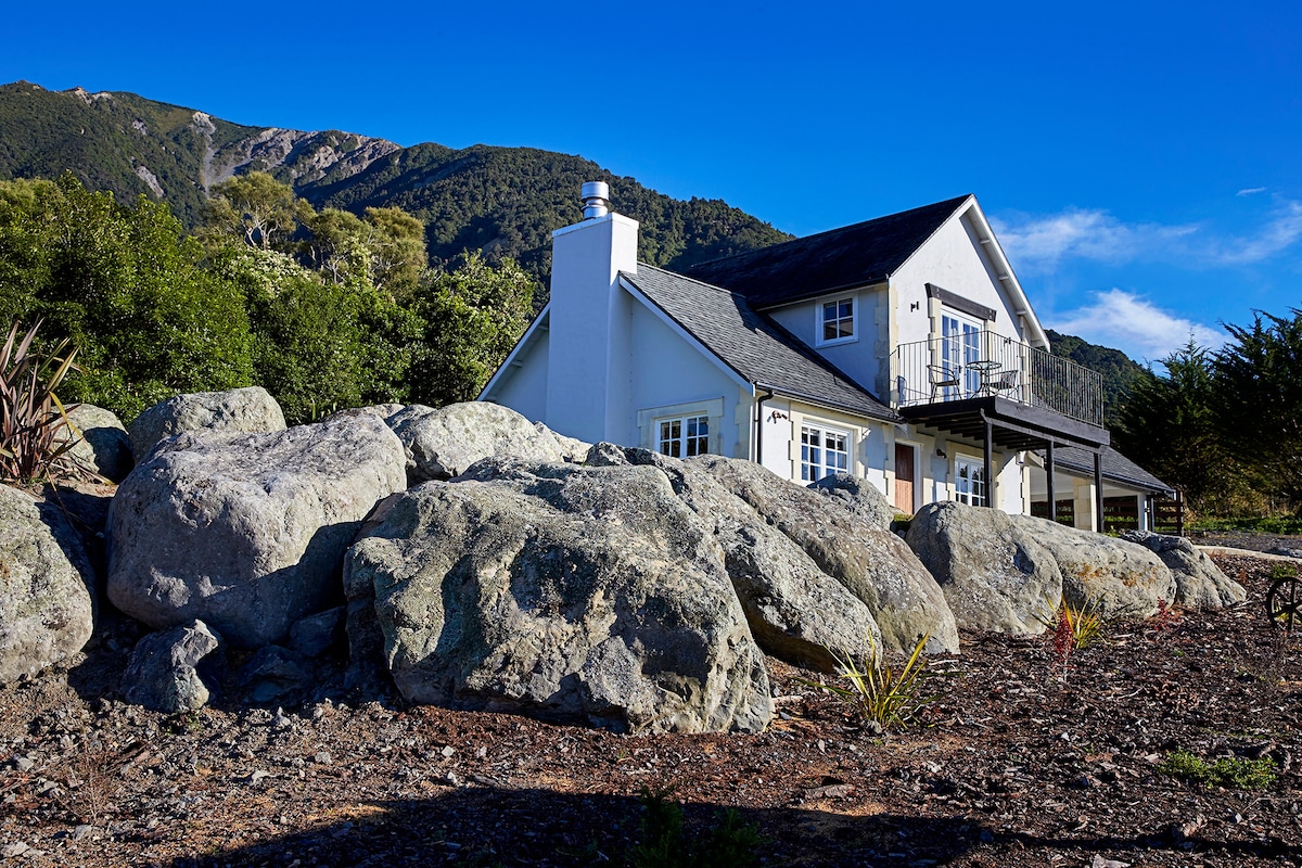 A rustic cottage is set against a backdrop of rolling hills and a rocky landscape. The two-story building features a balcony extending from the upper level, with large windows neatly arranged on the main façade. The surrounding natural greenery complements the peaceful setting.