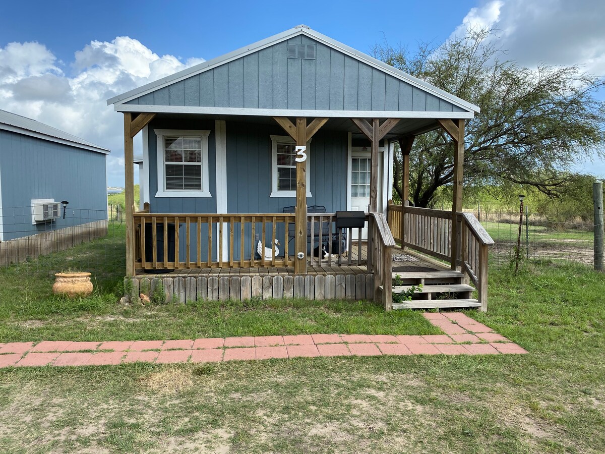 A blue cottage with a covered porch is shown. The porch is supported by wooden posts and features steps leading to the entrance. A barbecue grill is visible on the porch, and the surrounding area includes grass and a gravel pathway.