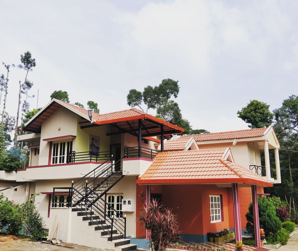 The exterior of a two-story house is presented, featuring a terracotta roof and a balcony. Lush greenery surrounds the property, contributing to a serene atmosphere. A flight of stairs leads to the entrance, enhancing accessibility and inviting exploration of the unique architecture.