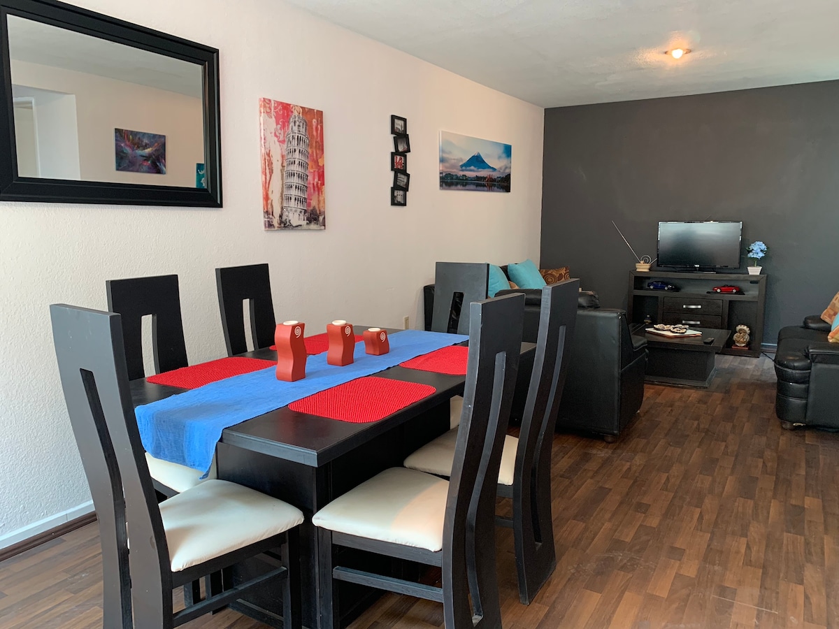 A dining area features a rectangular table set with a blue tablecloth, complemented by six chairs. A mirror hangs above the table, and colorful artwork decorates the walls. The living area includes a television unit and comfortable seating arrangements, all designed in a modern style.