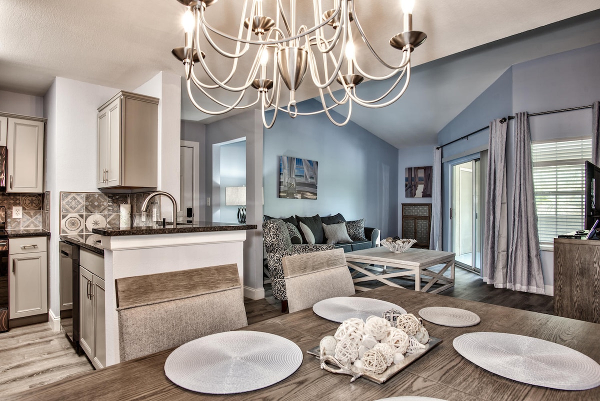An open-concept living and dining area is highlighted by a stylish chandelier above a modern dining table set with decorative plates. A spacious sofa is arranged around a coffee table, with large windows allowing natural light to brighten the room.