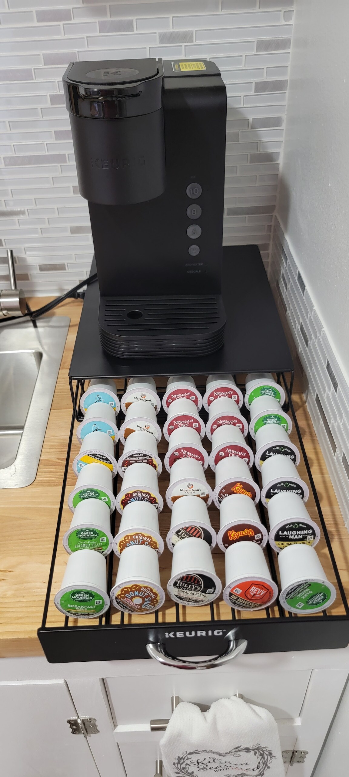 A coffee station is set up with a Keurig coffee maker positioned on a wooden countertop. Below, a tiered rack displays various coffee pods in a range of colors and flavors, providing easy access for brewing. A stainless steel sink is visible in the background.