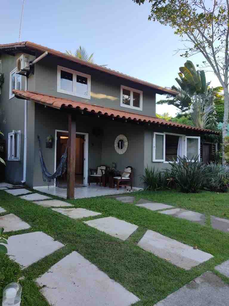 The exterior of the house showcases a two-story structure with a terra-cotta tiled roof. The entrance is framed by a small patio with two chairs and a hammock. Lush greenery and a pathway of stepping stones lead through the garden, enhancing the welcoming ambiance.