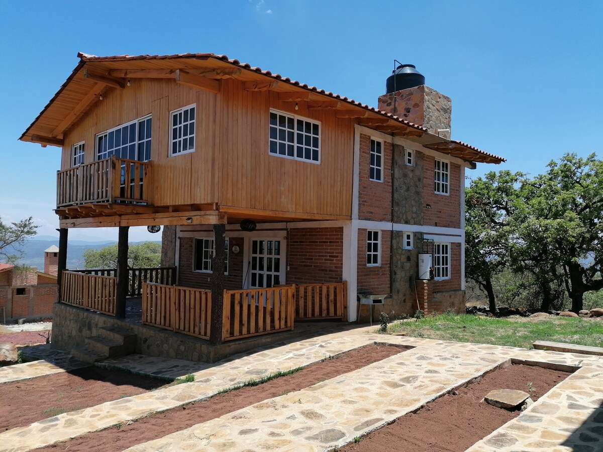 A two-story cabin featuring a wooden exterior with stone accents stands amidst a natural landscape. A spacious balcony is accessible from the upper floor, offering views of the surroundings. A stone pathway leads to the entrance, surrounded by earthy tones and greenery.