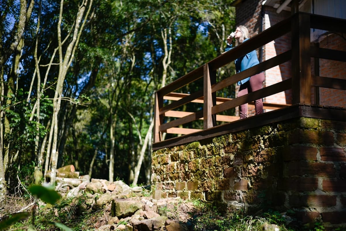 A wooden balcony extends from the house, surrounded by a lush native forest. The sturdy railing offers safety, while the stone foundation provides texture against the greenery. Sunlight filters through the trees, casting patterns on the ground, enhancing the natural setting.