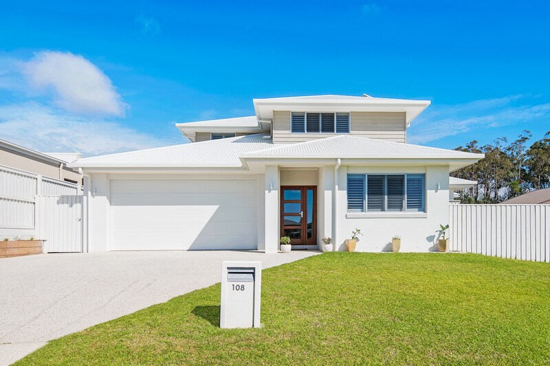 The modern two-story home features a clean white exterior with large windows and a welcoming front door. A manicured lawn and smooth driveway enhance the entrance, while decorative pots with plants are placed near the entrance, set against a clear blue sky.