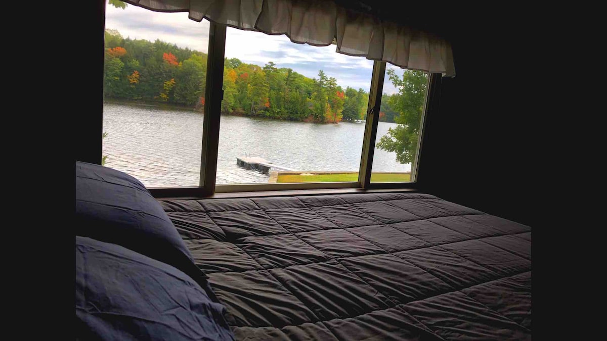 A spacious bed is positioned beside a large window, offering views of the serene water and surrounding trees. The scene showcases a private dock off the shore, while colorful autumn foliage enhances the natural landscape outside.