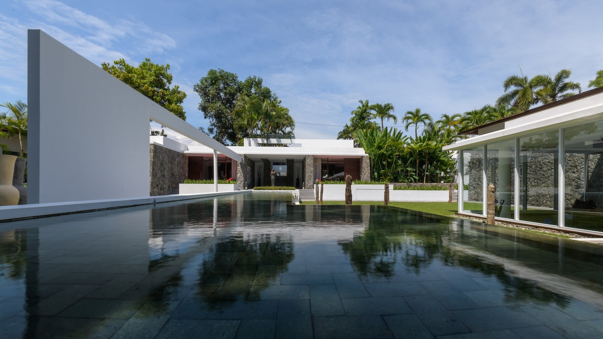 A modern architectural structure is reflected in the tranquil waters of the infinity pool. The surrounding landscape features lush tropical vegetation, with palm trees positioned in the background. The sleek design includes open spaces and large glass windows, inviting natural light into the villa.