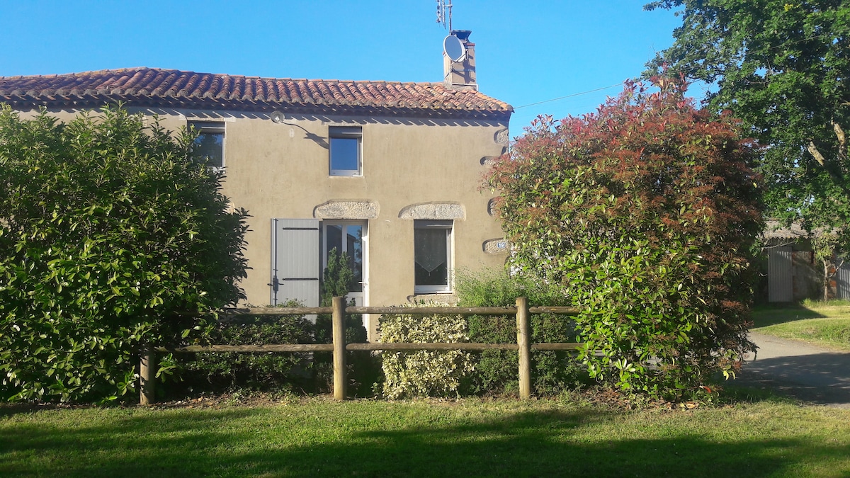 A charming house is surrounded by greenery, featuring a rustic façade with window shutters. The sloped roof tiles and chimney add character. A wooden fence delineates the property, and a variety of shrubs and trees are visible in the well-maintained garden.