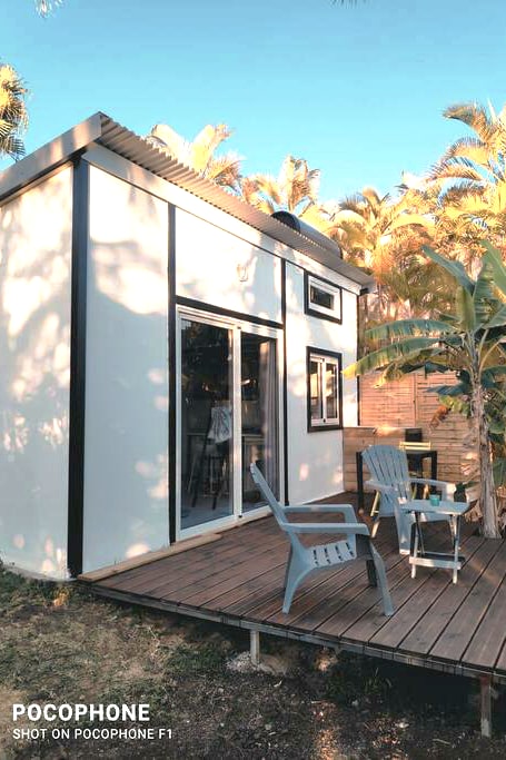 A modern bungalow is depicted with a sleek exterior and large glass doors. A small wooden deck features two blue chairs, inviting relaxation. Lush greenery, including palm trees, frames the structure, contributing to a tranquil outdoor setting.