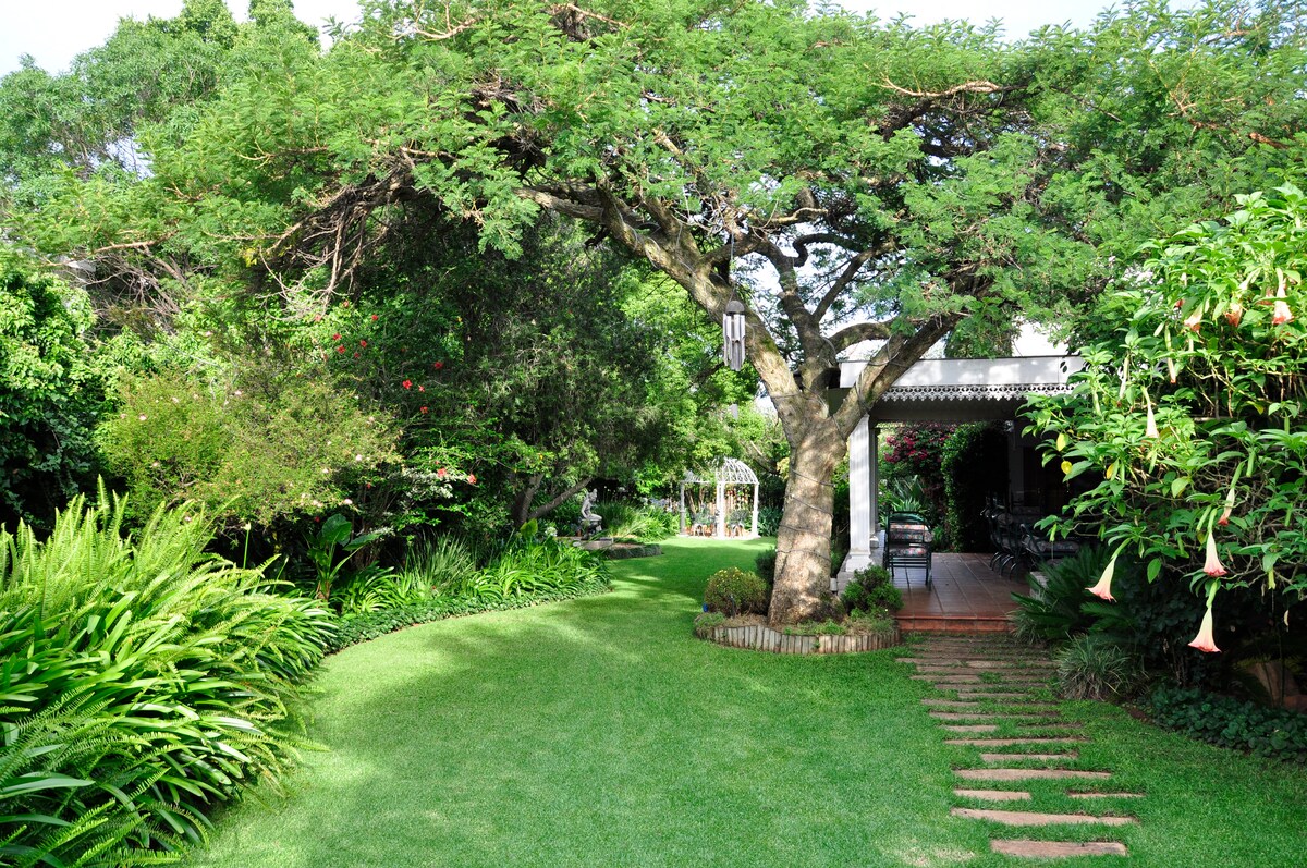 A lush garden is depicted, featuring a large tree and well-maintained grass. Stone stepping stones lead through the greenery, providing access to a shaded outdoor seating area partially visible in the background, surrounded by vibrant foliage and colorful flowers.