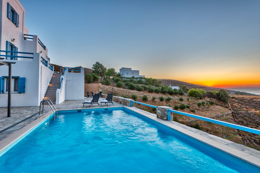 A clear blue pool is framed by a stone deck, where several lounge chairs are positioned for relaxation. Soft hues of sunset illuminate the landscape, highlighting gentle hills in the background. The modern architecture of the villa is visible beyond the pool area.