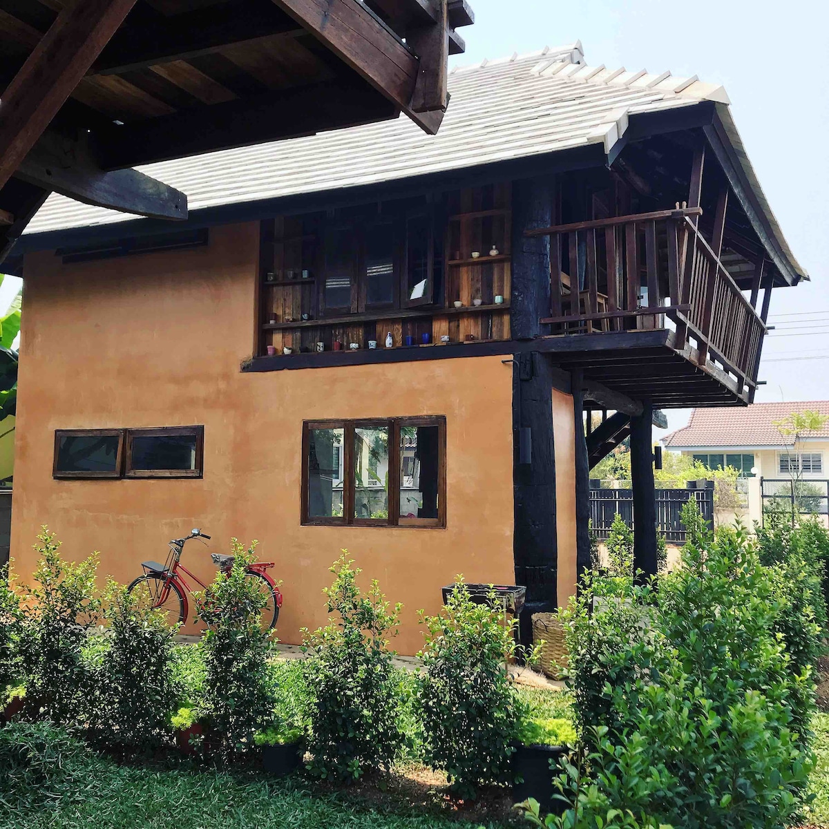 A charming Teak House is depicted with a rustic exterior featuring warm earthy tones. A balcony overlooks a well-maintained garden, while windows showcase the craftsmanship of the structure. A bicycle rests nearby, contributing to the inviting atmosphere of the property.