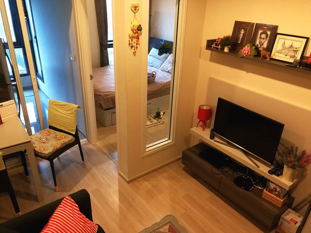 A modern living space is depicted, featuring a television mounted on a low shelf. A dining table with a multi-colored chair is visible next to a mirror, leading to a separated bedroom area with a neatly made bed. Natural light enhances the room's ambiance.