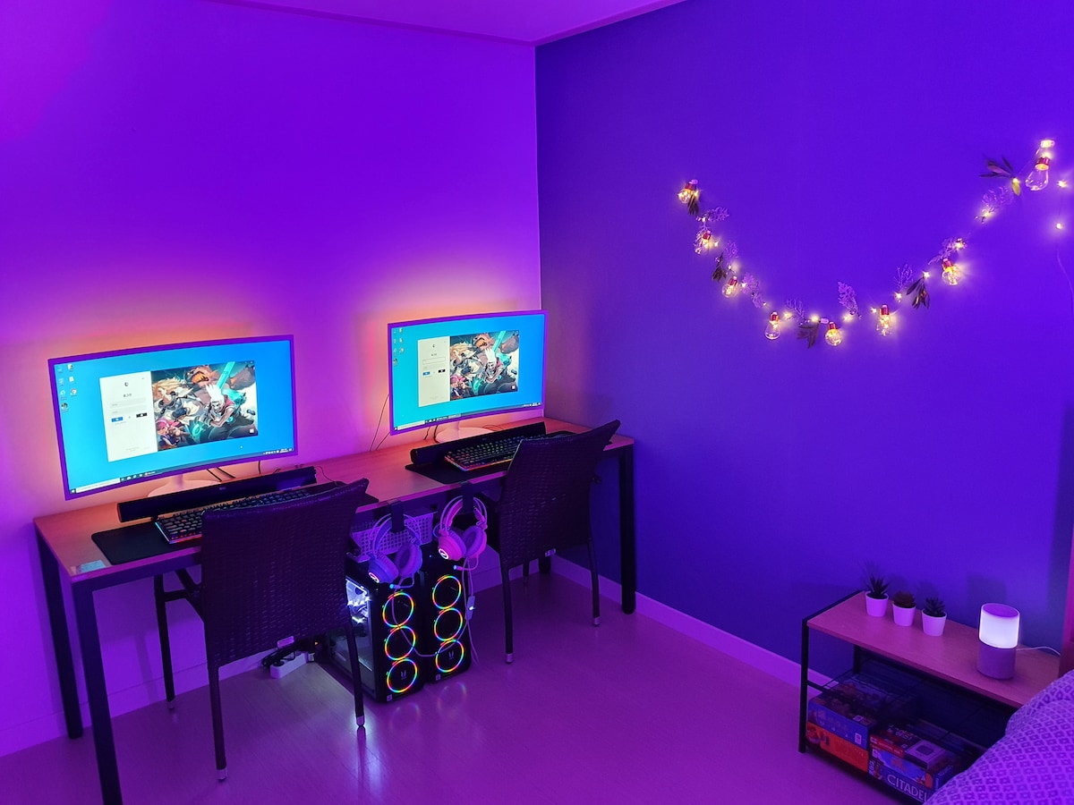 A dedicated gaming area is presented with two monitors illuminating the space in soft blue and purple tones. Each computer is set on a simple desk, accompanied by comfortable chairs. Decorative lights adorn the wall, enhancing the modern atmosphere.