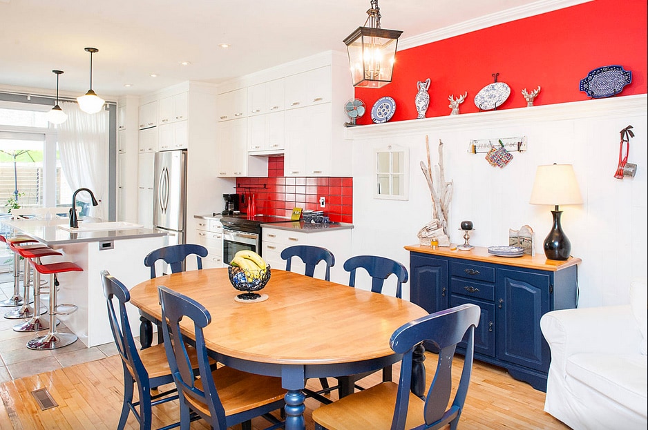 An open kitchen and dining area showcases a wooden dining table surrounded by blue chairs. A large island with seating sits nearby, complemented by modern appliances and white cabinetry. Bright red accents and decorative plates enhance the overall charm of the space.