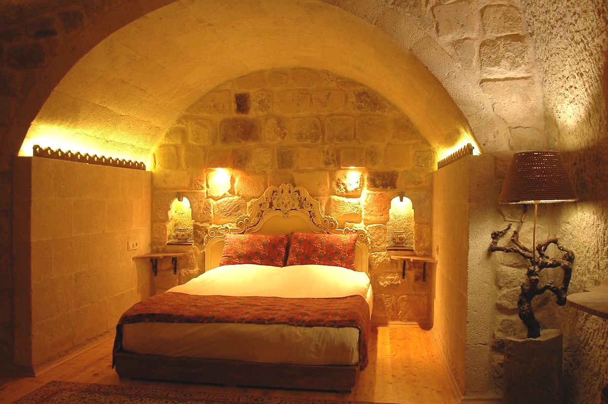 A cave-style living area features a spacious bed with a patterned blanket, flanked by softly glowing sconces. Stone walls provide rustic charm, with a wooden floor and decorative lamp enhancing the ambiance. An overall sense of warmth is conveyed by the gentle lighting.