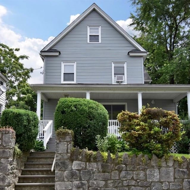A charming cottage exterior features a grey wooden facade set against a clear blue sky. A stone pathway leads to a front porch, flanked by neatly trimmed shrubs and vibrant greenery. The inviting entrance is illuminated by natural sunlight, suggesting a welcoming atmosphere.