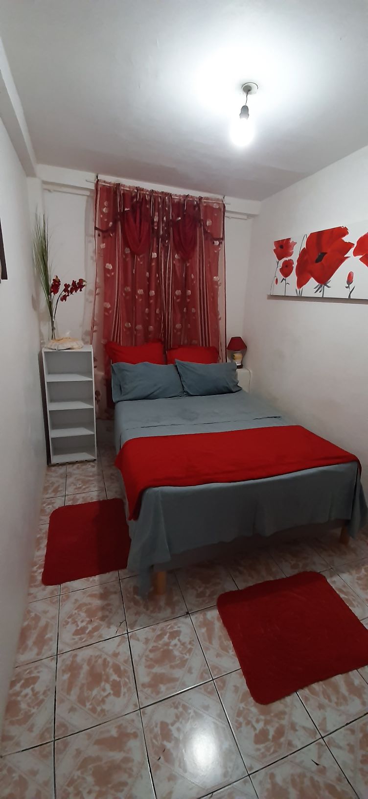 A cozy bedroom is showcased with a comfortable bed dressed in gray and red linens. A floral wall art piece complements the decor, while red rugs add a touch of color. A small bedside table and a chest of drawers provide functional storage.