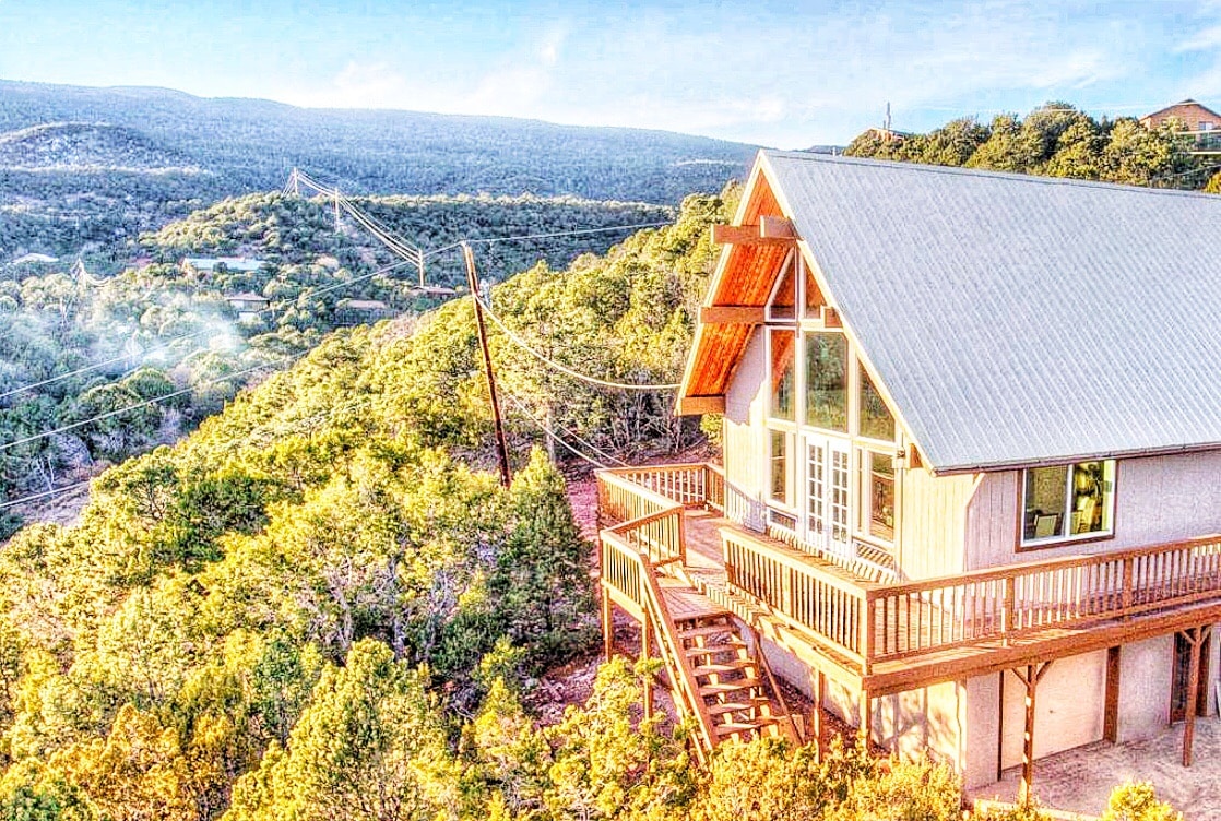 The A-frame home is nestled among greenery, featuring a spacious deck with a railing that overlooks the surrounding hills. Large windows allow for ample natural light, and a staircase leads down to the lower level, enhancing the connection with the natural landscape.