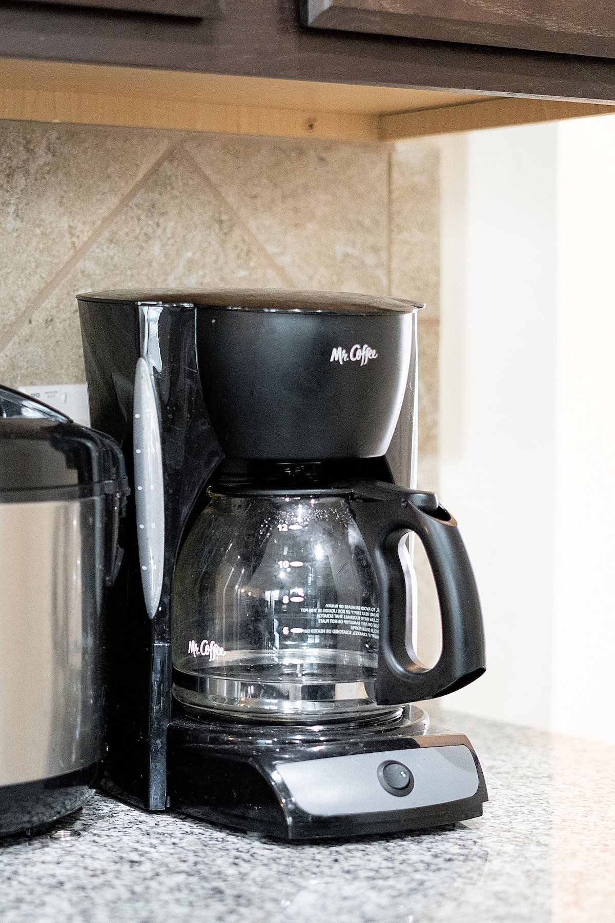 A coffee maker is positioned on a granite countertop, featuring a glass carafe and black plastic exterior. The appliance includes measurement markings on the side and a single control button at the base.