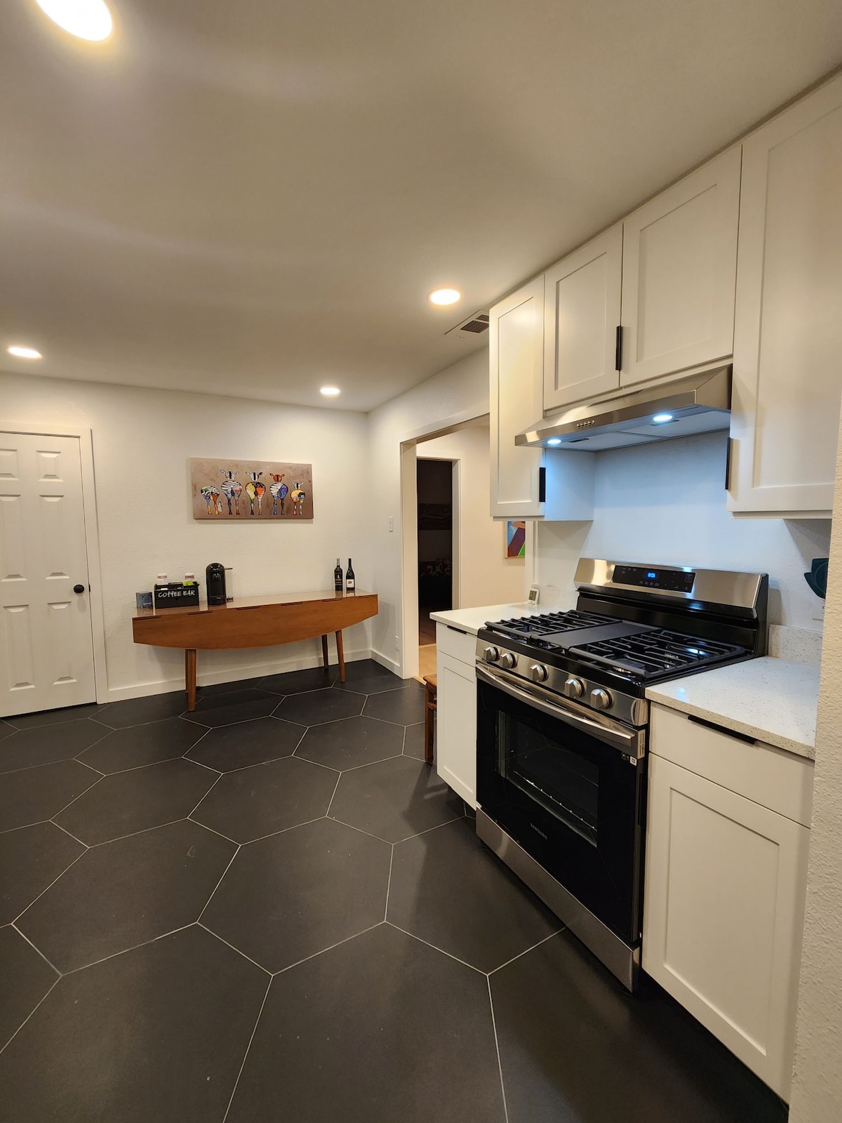 The kitchen displays modern white cabinetry, a stainless-steel gas range, and sleek countertops. A wooden table with bottles of wine is positioned against the wall. Black hexagonal tiles cover the floor, enhancing the contemporary aesthetic.