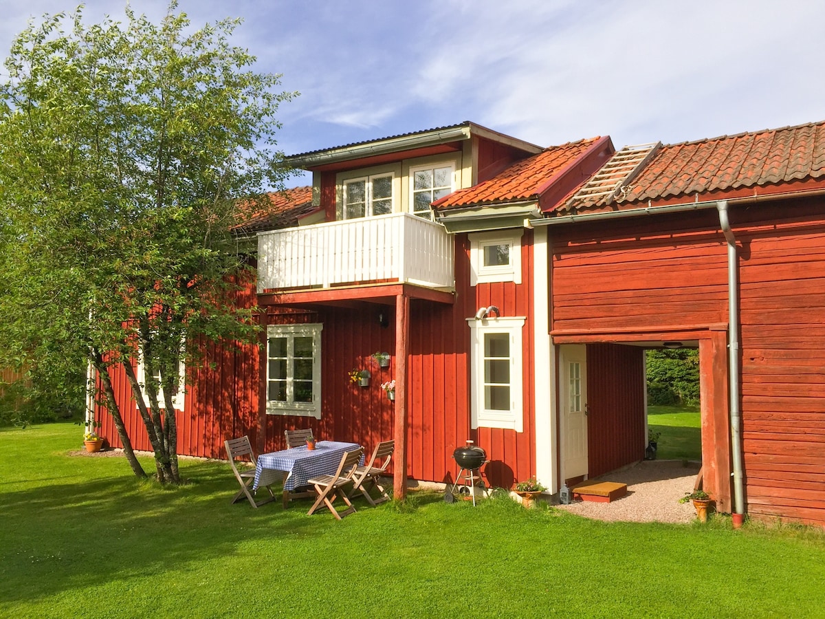 The exterior of a charming red house is showcased, featuring a white balcony above a gravel path. Wooden folding chairs and a table are set on the lush green lawn, surrounded by a tree. A barbecue grill is positioned nearby.