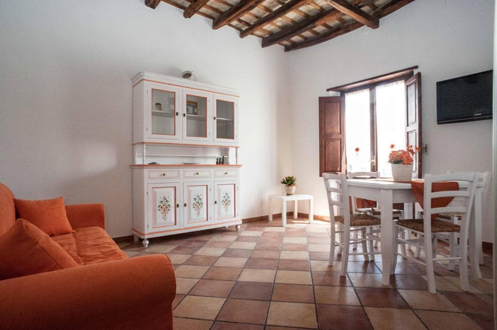 A bright living area features a comfortable orange sofa and a white dining table surrounded by wooden chairs. A decorative cabinet adds charm, while natural light streams through the window, highlighting the tiled floor and wooden ceiling beams.
