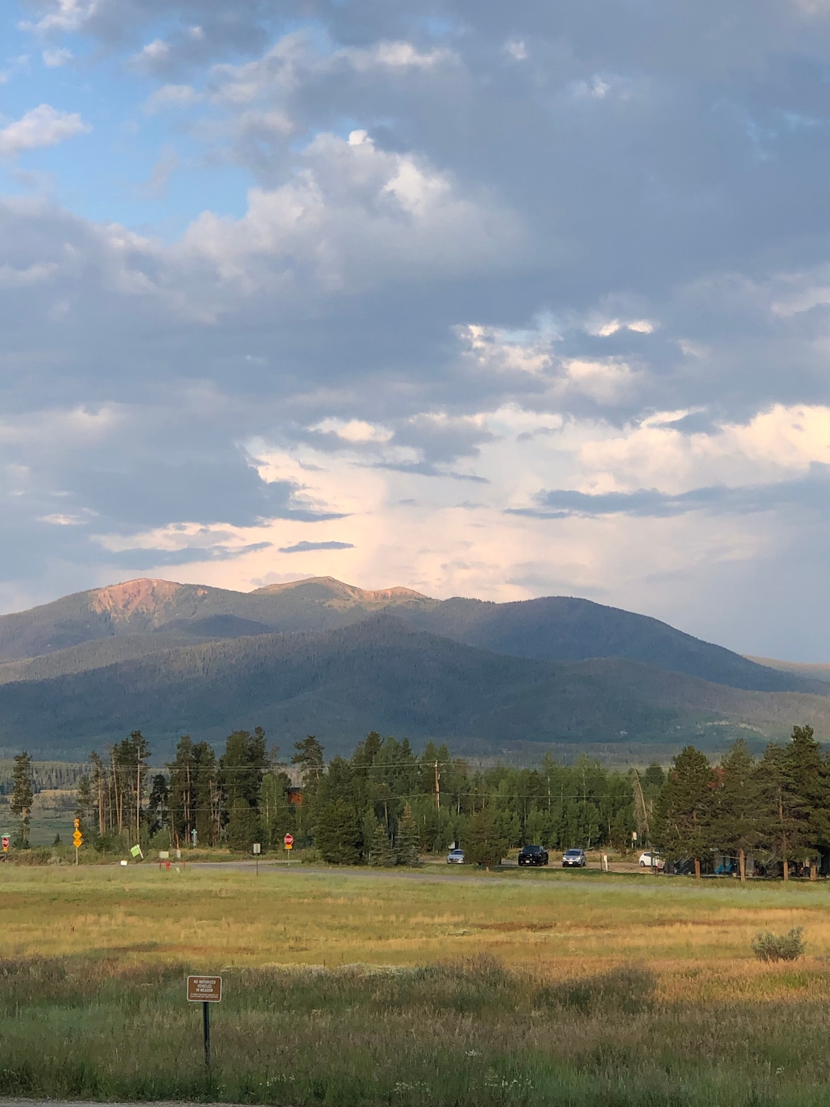 A panoramic view of majestic mountains is captured, showcasing varying shades of green forest and rugged terrain. Fluffy clouds drift across a blue sky, casting soft shadows on the peaks, highlighting their natural beauty.