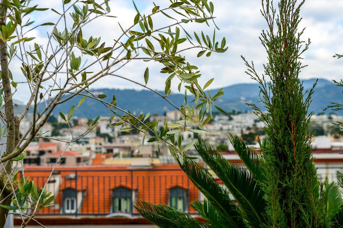 A view from the balcony showcases rooftops dotted with greenery and terracotta tiles, framed by lush plants. Soft hills rise in the background, with clouds gently drifting across the sky, offering a sense of tranquility in the urban landscape.