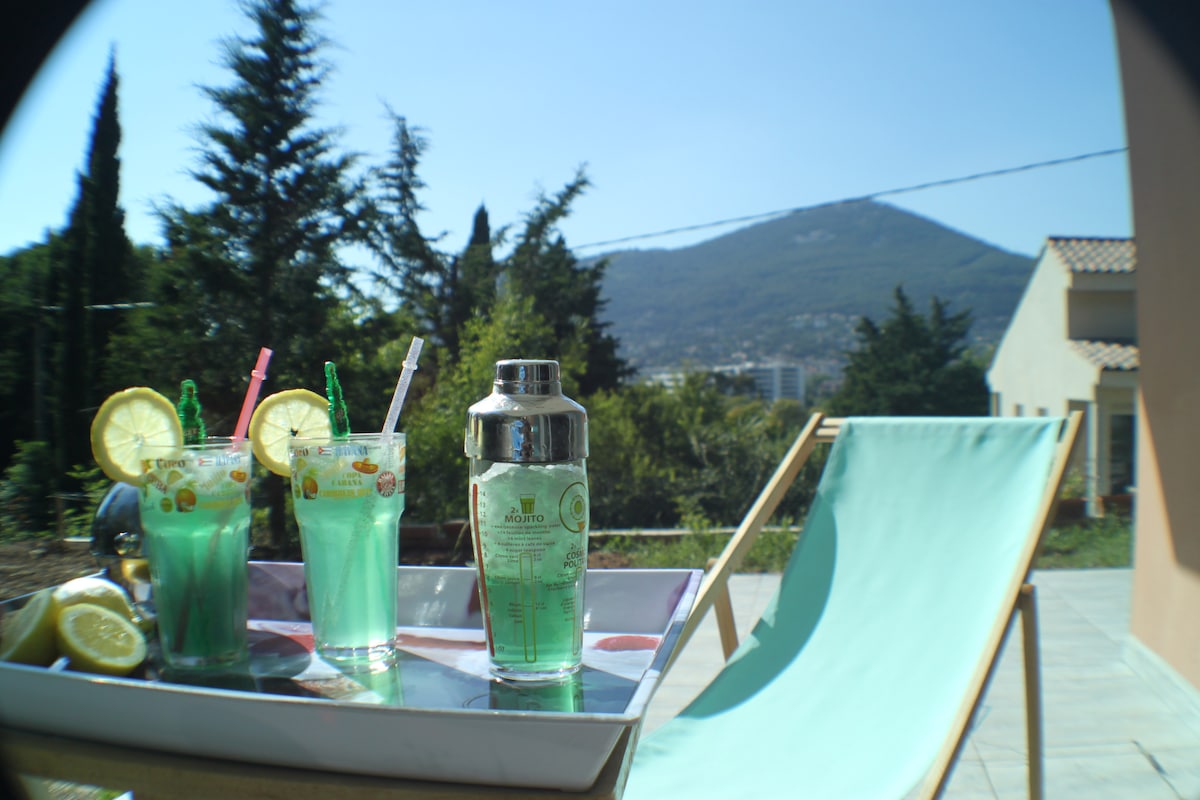 A tray holds two vibrant drinks garnished with lemon slices, accompanied by a cocktail shaker. A turquoise lounge chair is positioned nearby under clear blue skies, with a backdrop of lush greenery and a mountain view.
