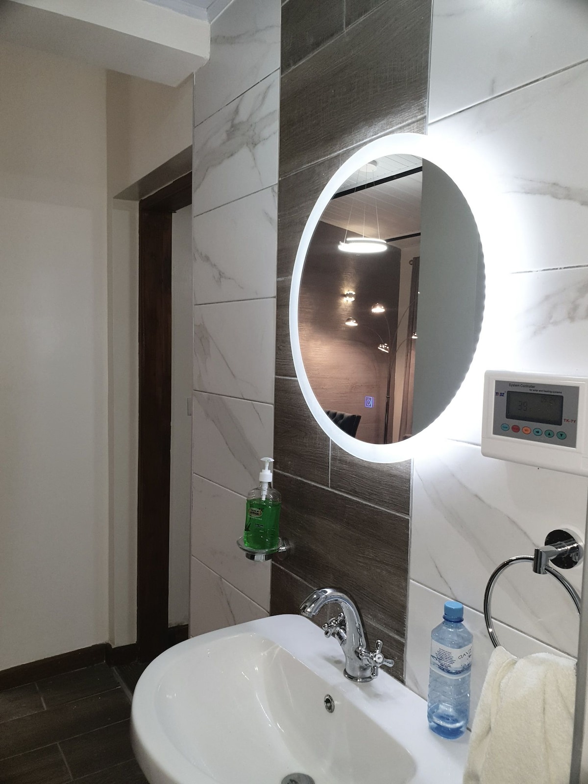 A modern bathroom features a circular mirror illuminated by soft light. A sleek sink sits below the mirror, accompanied by a chrome faucet. Nearby, a hand soap dispenser and a bottle of water are present, with textured tiles adding a stylish touch to the walls.