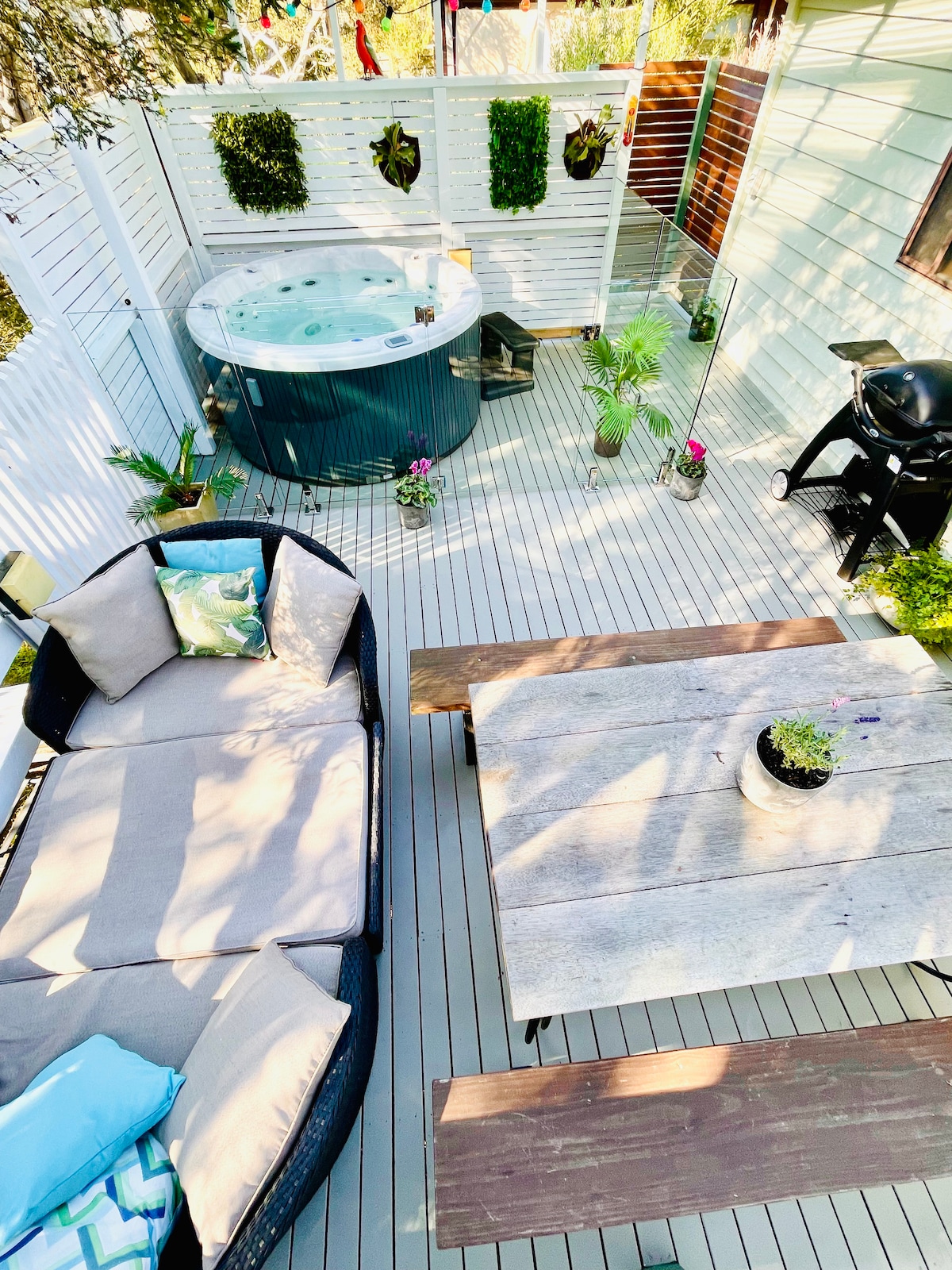 An outdoor deck area features a hot tub, surrounded by greenery and decorative plants. A wooden table and benches are highlighted, with a charcoal grill nearby. Cozy seating is arranged with plush cushions, creating a relaxing space for enjoyment.