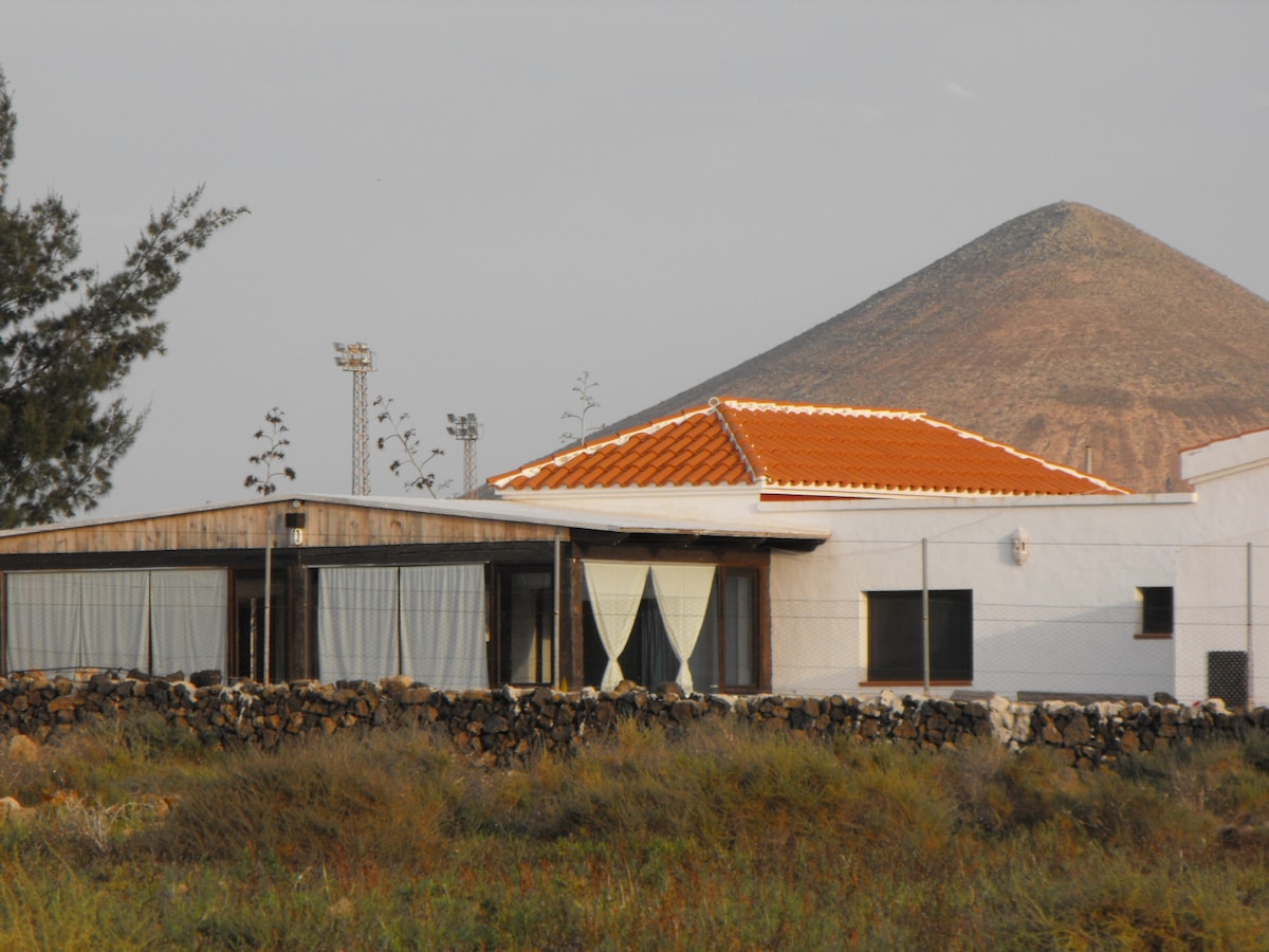 The exterior view of the home is presented, featuring a terracotta-tiled roof and wooden accents. A stone wall surrounds the property, and large windows allow for ample natural light. In the background, a smooth volcanic mountain rises, accentuating the serene landscape.