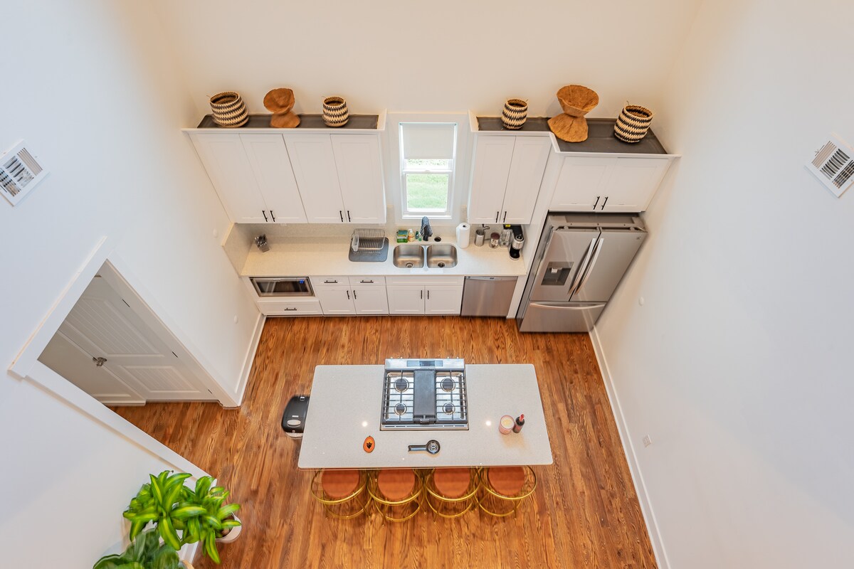 The kitchen is designed with a central island featuring a gas cooktop and bar seating. Light wood flooring complements the modern cabinetry and stainless-steel appliances. Natural light floods the space through large windows, enhancing the clean lines and open layout.