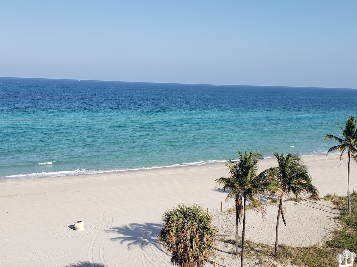 A serene view of the ocean stretches across the image, showcasing vibrant turquoise waters merging into deeper blues. The sandy beach is bordered by palm trees, creating a peaceful atmosphere. Clear skies enhance the tranquil scenery, inviting relaxation by the seaside.