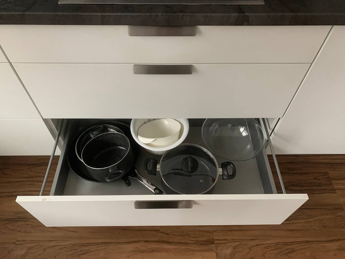 A kitchen drawer is displayed, containing a variety of cookware including pots and a glass lid. A white bowl and a small pan are also visible, all neatly arranged on a grey liner, which complements the sleek white cabinetry.