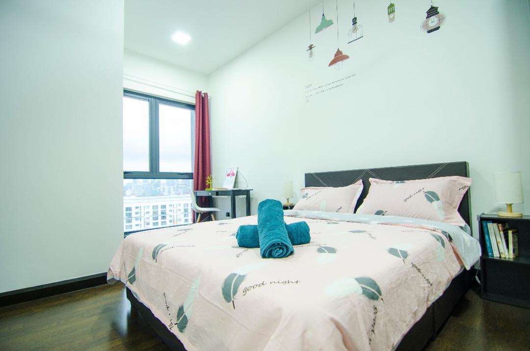 A spacious sleeping area is presented with a queen-size bed adorned with a pink and patterned bedspread. Two bedside lamps provide soft lighting beside the bed. A writing desk with a chair is positioned near a large window, offering a glimpse of the city skyline.
