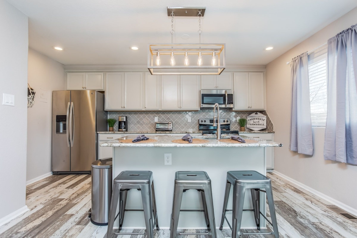 A modern kitchen features a large island with three metal bar stools. Stainless steel appliances and light cabinets complement the marble countertop. A chandelier hangs above the island, providing illumination, while soft curtains frame the window, allowing natural light to enter.
