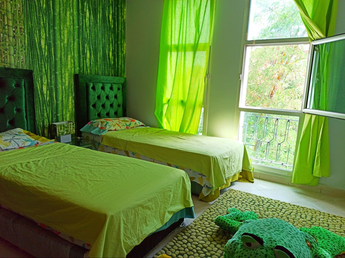 A bright, vibrant bedroom features two spacious beds topped with colorful linens. The walls are adorned in various shades of green, complemented by matching curtains flowing along the windows. A playful frog-shaped rug sits on the floor, enhancing the room's cheerful atmosphere.