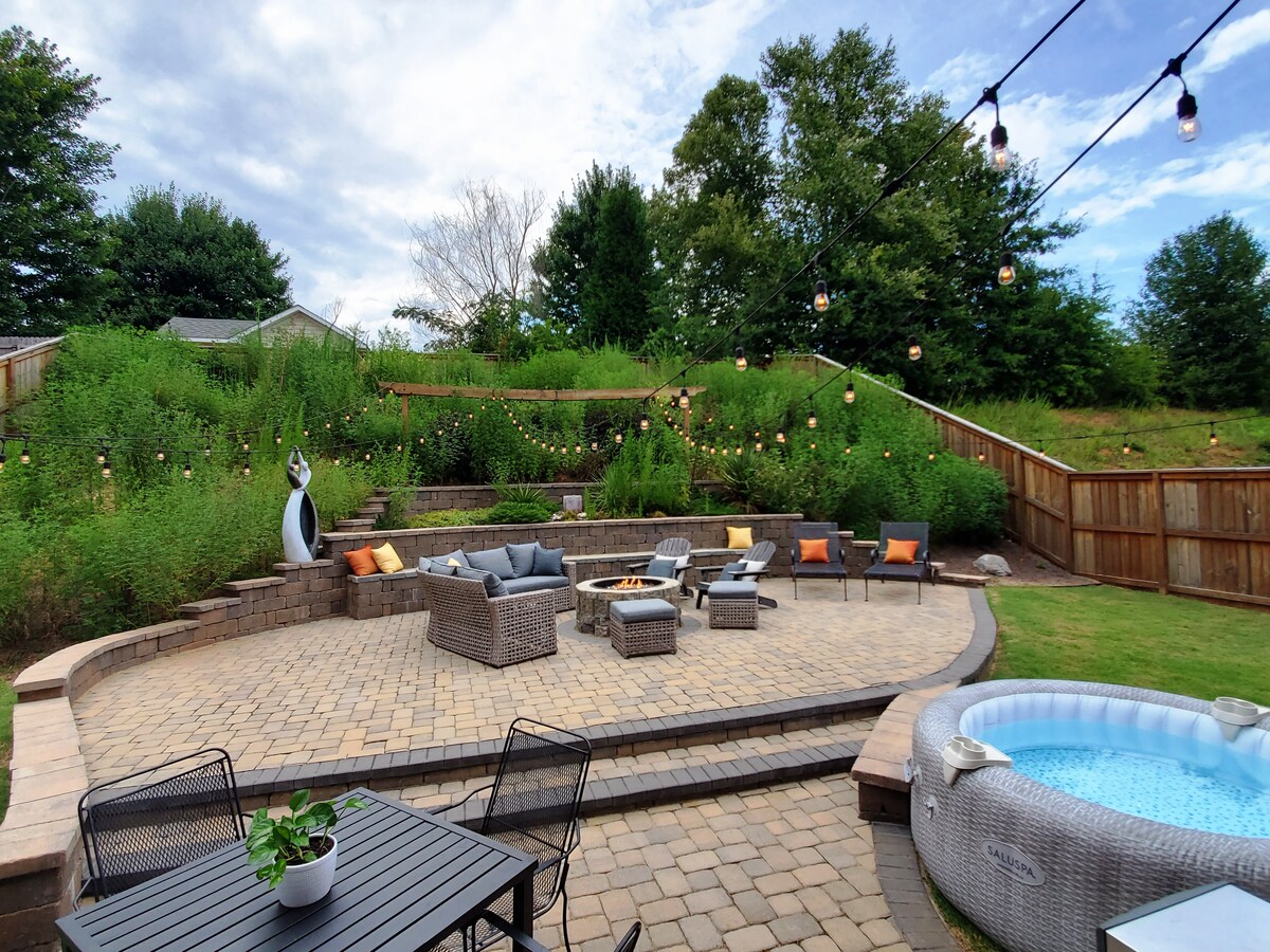An outdoor seating area features stone pavers with a circular arrangement of cushioned chairs and a fire pit at the center. String lights hang above, and a hot tub is positioned nearby, surrounded by lush greenery and terraced hills.