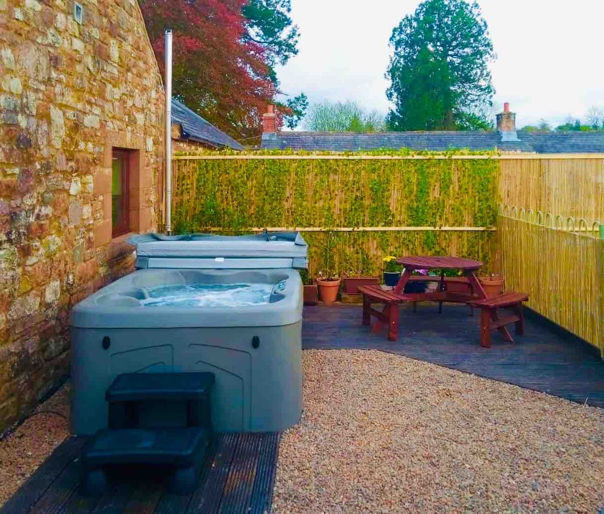 An outdoor area features two hot tubs surrounded by natural stone walls and greenery. A picnic table is positioned on the decking, complemented by planters. Gravel flooring creates a relaxed atmosphere in this private courtyard setting.