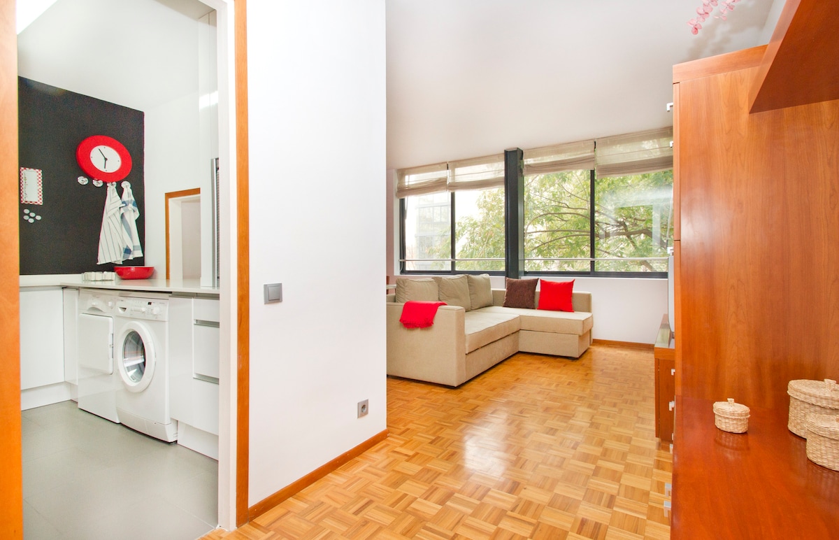 The image showcases a bright living area featuring a large beige sectional sofa with red accents. A sliding glass door allows natural light to fill the space, while a light wood floor creates a warm atmosphere. A separate laundry area is visible through an open door.